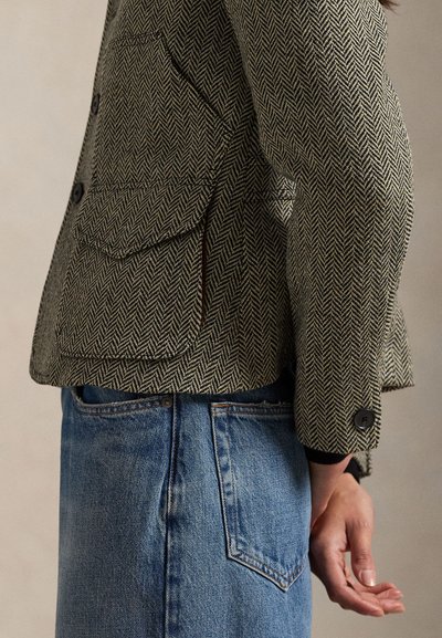 Kurtka w jodełkę w kolorze szaro-beżowym, z dużymi przednimi kieszeniami, guzikami i dopasowanym krojem, noszona na niebieskich dżinsach.