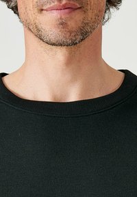 Maglione in cotone nero a girocollo con scollatura a costine, texture liscia e silhouette aderente. Pelle con una leggera peluria visibile all'altezza del collo.