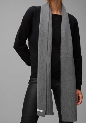Personne portant une longue écharpe en tricot grise sur un pull noir et un pantalon en cuir noir, debout devant un fond gris uni.