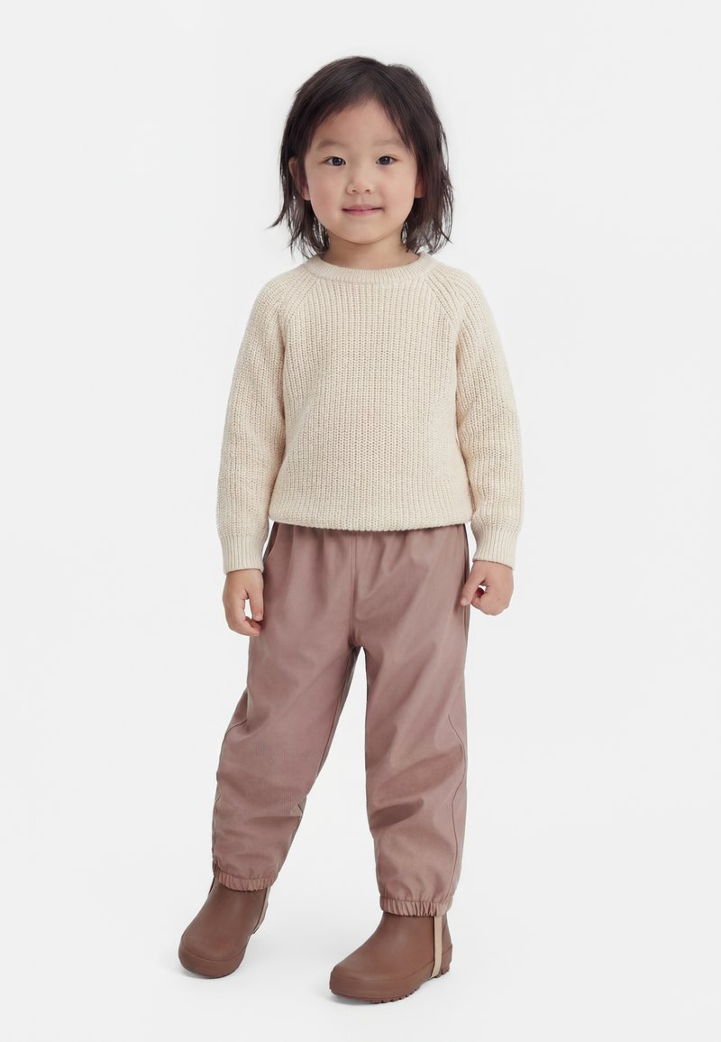 Malo dijete stoji, nosi kremasto pleteni džemper, praškasto roza hlače s elastičnim manšetama i smeđe cipele na navlačenje na običnoj bijeloj pozadini.