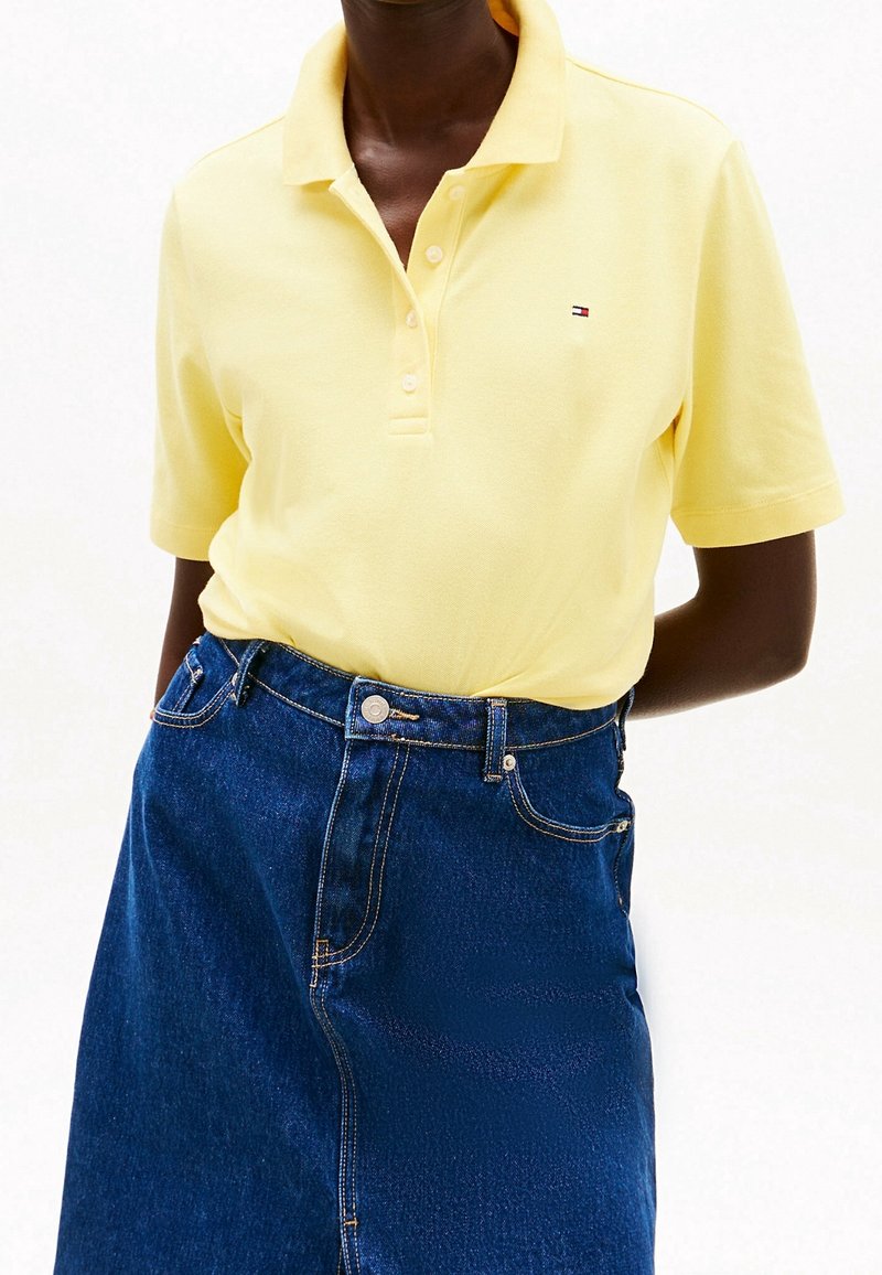 Personne portant un polo à manches courtes jaune pâle, rentré dans une jupe en denim bleu foncé taille haute avec boutons à l'avant et poches.