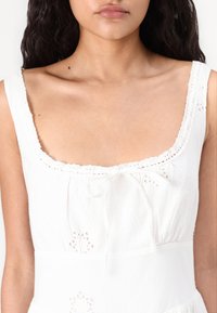 Abito bianco con scollatura quadrata, bordo in pizzo e ricami floreali. Tessuto leggero e corsetto aderente con dettaglio di lacci sul davanti.