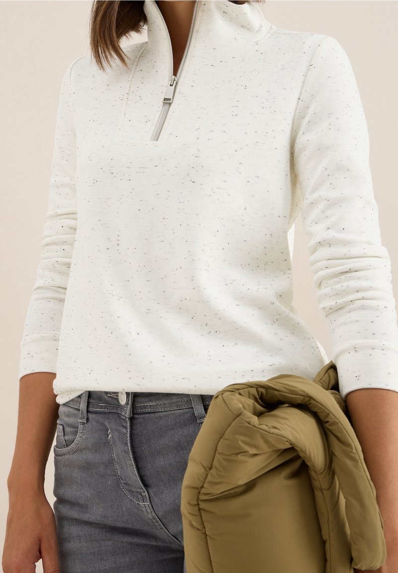 Personne portant un pull à fermeture éclair quatre-quarts blanc tacheté et un jean gris, tenant une veste matelassée vert olive dans une main.
