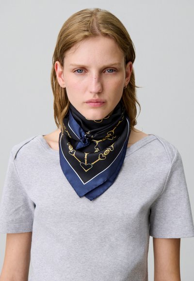 Bufanda de seda con un fondo azul marino y un patrón de corazones y cadenas doradas, doblada en forma de triángulo y llevada alrededor del cuello.