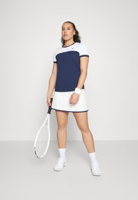 Ναυτικό και λευκό αθλητικό σετ που περιλαμβάνει ένα εφαρμοστό τοπ με σχέδιο χρωματικής μπλοκ και μια ταιριαστή φούστα. Περιλαμβάνει επίσης μια ρακέτα τένις και αθλητικές ταινίες για τους καρπούς.