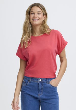 Naeratav naine, kellel on pikad blondid juuksed, kannab korallikarva lühikeste varrukatega T-särki ja siniseid teksaseid, üks käsi taskus, ühevärvilise tausta ees.