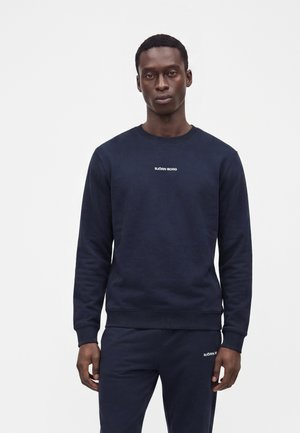 Homme portant un sweatshirt bleu marine et un pantalon assorti avec le logo "Björn Borg" sur la poitrine et la cuisse, debout devant un fond blanc uni.