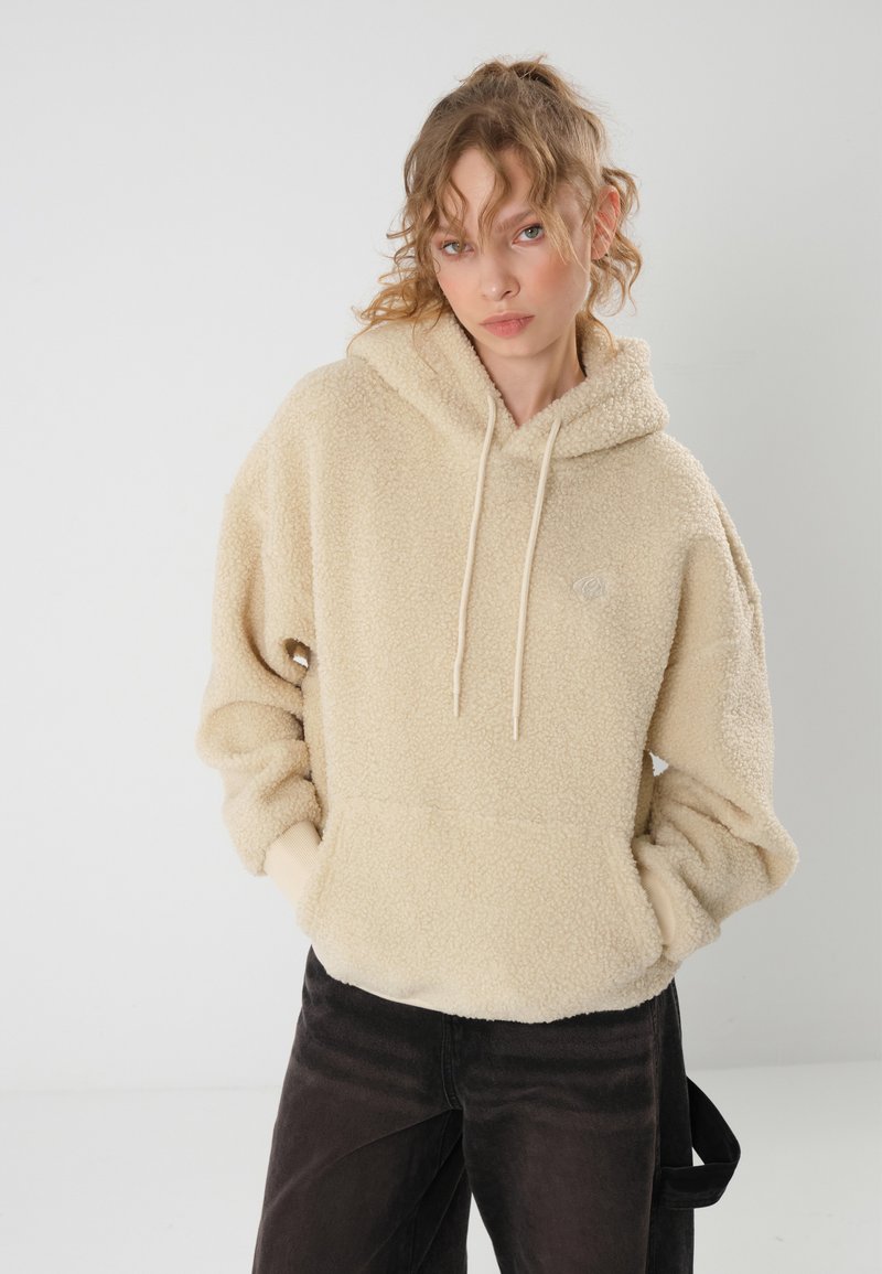 Felpa beige, morbida, con cappuccio regolabile e tasca frontale, realizzata in materiale peloso. Abbinata a pantaloni neri a gamba larga.
