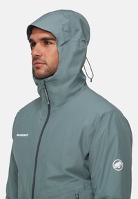 Vattentålig, grå Mammut-jacka med en strukturerad huva, framficka med dragkedja och logotyp på vänster ärm. Slätt, lättviktsmaterial.