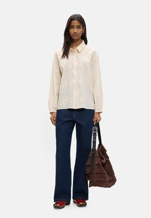 Kobieta ubrana w kremową, kwiatowo haftowaną bluzkę, ciemne szerokie jeansy, czerwone sandały, trzymająca dużą brązową zamszową torbę z falbankami.