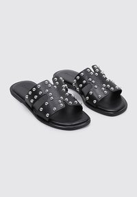 Chanclas negras con dos correas anchas adornadas con tachuelas plateadas. Suela plana con una textura suave y un diseño de punta abierta.
