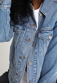 Light blue denim jacket with button closure, two chest pockets, and a relaxed fit. Features faded texture and visible stitching details.
