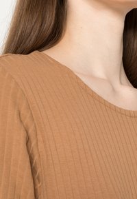 Brun ribbad topp med rund halsringning, tillverkad av mjukt tyg med en subtil textur, som visar en del av axeln och nacken.