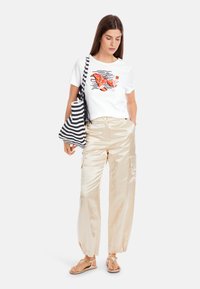 Weißes Grafik-T-Shirt mit einem Design von Hummer und Cocktail, kombiniert mit glänzenden, hellbeigen, weit geschnittenen Hosen und einer schwarzen gestreiften Tragetasche.