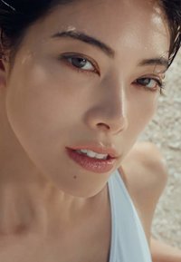 Gros plan sur une personne avec une peau claire et des cheveux légèrement humides. Ses traits incluent des yeux brun clair et une couleur de lèvres naturelle. Un vêtement blanc est visible.