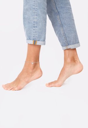 Cavigliera d'argento con un ciondolo rotondo, indossata su un piede nudo. Il piede è posizionato accanto a dei jeans arrotolatiazzurri chiari con cuciture arancioni.