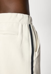 Cremefarvede sweatpants med en struktureret stof, der har sidelommer og sorte accentstriber langs siderne. Elastisk talje.