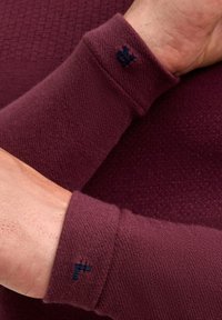 Bordowy sweter z teksturowanymi rękawami o prążkowanym wzorze i małym granatowym logo na mankiecie. Miękka, dzianinowa tkanina.