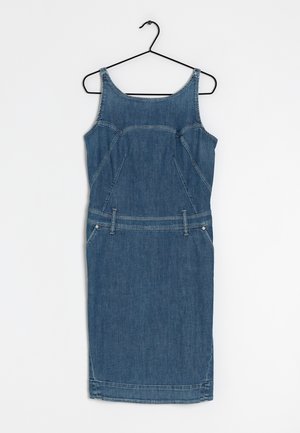 Vestido sin mangas de mezclilla azul hasta la rodilla con trabillas para cinturón, colgado en una percha de alambre negra sobre un fondo blanco liso.