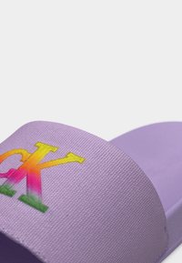 Παντόφλα πλατφόρμας σε λιλά χρώμα με υφασμάτινο λουρί που φέρει πολύχρωμο βαθμιδωτό λογότυπο "CK" σε απλό λευκό φόντο.