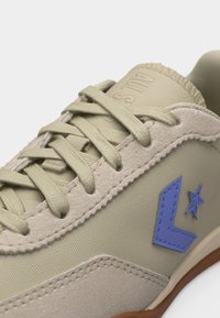 Μπεζ αθλητικό παπούτσι με υφασμάτινη υφή και λεπτομέρειες από σουέντ, μπεζ κορδόνια, μοβ βέλος και λογότυπο αστεριού, με καφέ άκρη σόλας ορατή.