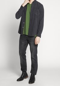 Mörkgrå skjorta med knappar över en grön v-ringad t-shirt, i kombination med mörka jeans och svarta läderstövlar vid ankeln. Enkel, avslappnad design.