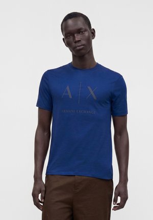 Homme portant un t-shirt bleu Armani Exchange et un pantalon marron, debout devant un fond clair uni.