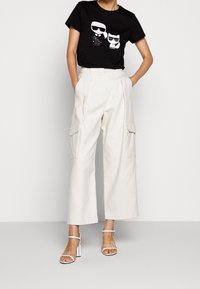 Camiseta gráfica negra con dos personajes de dibujos animados; combinada con pantalones cargo de pierna ancha en color beige y sandalias blancas con tiras.