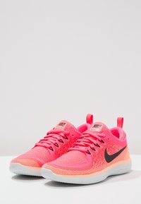 Une paire de chaussures de course Nike roses vives avec des logos swoosh noirs, des semelles blanches et des languettes sur une surface blanche.