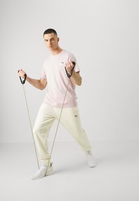 Camiseta de algodón rosa con el logo de PUMA, pantalones de chándal crema y zapatillas blancas. El modelo sostiene bandas de ejercicio en una pose dinámica. Fondo liso.