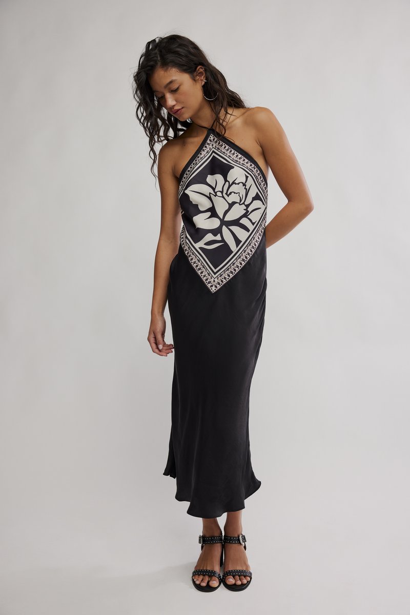 Γυναίκα με μαύρο φόρεμα halter neck με μεγάλο λευκό λουλουδάτο σχέδιο μπροστά, φορώντας μαύρα σανδάλια με τρουκς, ποζάροντας μπροστά σε ουδέτερο φόντο.