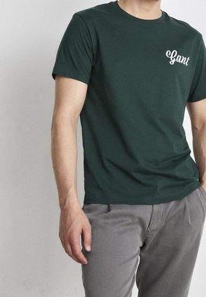 Mężczyzna ubrany w ciemnozieloną koszulkę z krótkim rękawem z logo "Gant" na piersi i jasnoszare spodnie, stojący na jednolitym tle.