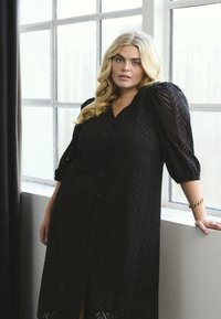 Robe noire présentant un motif texturé, un devant à boutons et des manches bouffantes. Le tissu a une apparence lacée avec une fente sur l'ourlet.