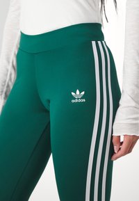 Green athletic leggings with a high waistband, featuring three white vertical stripes on the side and a small white Adidas logo.