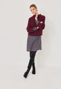 Weinrot gestrickter Pullover mit gerippter Textur, über einem gemusterten dunklen Kleid getragen. Schwarze Stiefel bis zur Oberschenkelhöhe runden das Outfit ab.
