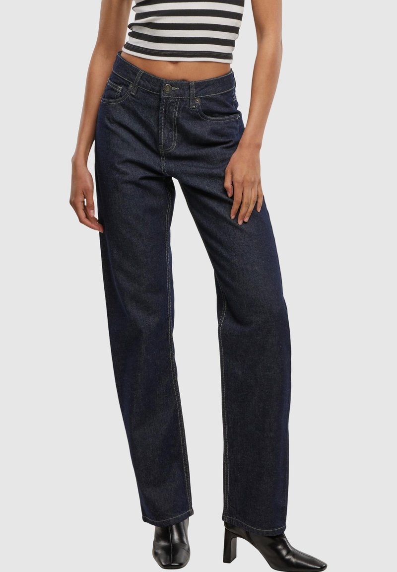 Femme portant un jean droit bleu foncé taille haute, des bottines noires à bout carré et talons, et un top court rayé noir et blanc.