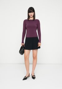 Donna che indossa un top a maniche lunghe viola, una gonna plissettata nera, tacchi a punta neri, mentre tiene una borsa nera su uno sfondo bianco uniforme.