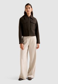 Bruine cropped jas met een rits en twee voorzakken, gecombineerd met crèmekleurige wijde broek en donkerbruine schoenen. Neutrale achtergrond.