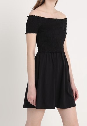 Jeune femme portant une robe noire épaules dénudées à manches courtes, avec un corsage ajusté texturé et une jupe évasée.