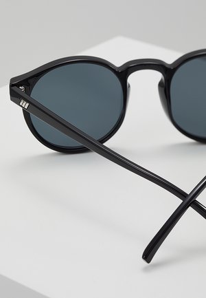 Sorte runde solbriller med mørke glas, der ligger på en hvid overflade, med tynde, blanke sorte stel og en lille sølvdetalje på hængslet.