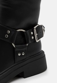 Stivale in pelle nera con tacco spesso, dotato di una cinghia decorativa con dettagli in metallo argento e accenti a forma di cerchio. Texture liscia, design robusto.