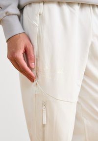 Crème-kleurige sportbroek met een strakke, gladde textuur, voorzien van een zijritszak en een reliëflogo, ontworpen voor een moderne pasvorm.