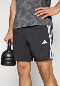 Mustad spordipüksid valgete küljeribade ja Adidas'i logoga. Modell hoiab ühes käes musta raskust. Tekstureeritud kangas.