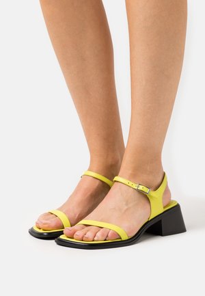 Gelbe Leder-Sandaletten mit Blockabsatz, zwei dünnen Riemen über den Zehen und einem Knöchelriemen mit silberner Schnalle. Schwarze Sohle.