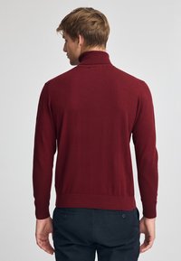 Stickad bordeauxtröja med hög krage, långa ärmar och ribbad fåll. Mjuk textur, figurnära design, minimalistisk stil.