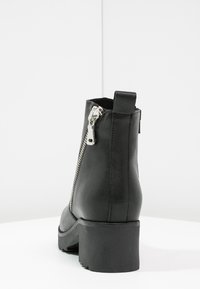 Bottine noire en cuir avec fermeture éclair latérale, talon épais et surface texturée. Comprend une languette à l'arrière pour un enfilage facile.