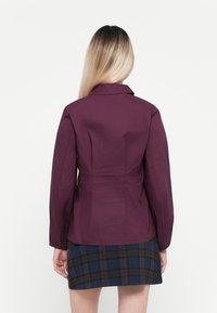 Bordeauxkleurige fitted blazer met kraag en lange mouwen, gecombineerd met een geruite rok. Gladde textuur, minimaal ontwerp en geen zichtbare hardware.