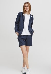 Felpa blu navy con cappuccio, tasche frontali e dettagli in cerniera bianca abbinata a pantaloni corti blu navy. T-shirt bianca sotto.