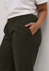 Pantalons décontractés vert foncé en tissu doux, dotés d'une taille élastique et de poches latérales. Portés avec un t-shirt blanc.
