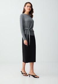 Femme portant un haut gris à manches longues avec un lien sur le côté, une jupe midi noire et des sandales noires à talons, debout devant un fond clair uni.